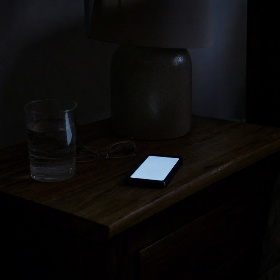 A dark bedside table at night lit only by a phone screen, beside a glass of water and reading glasses