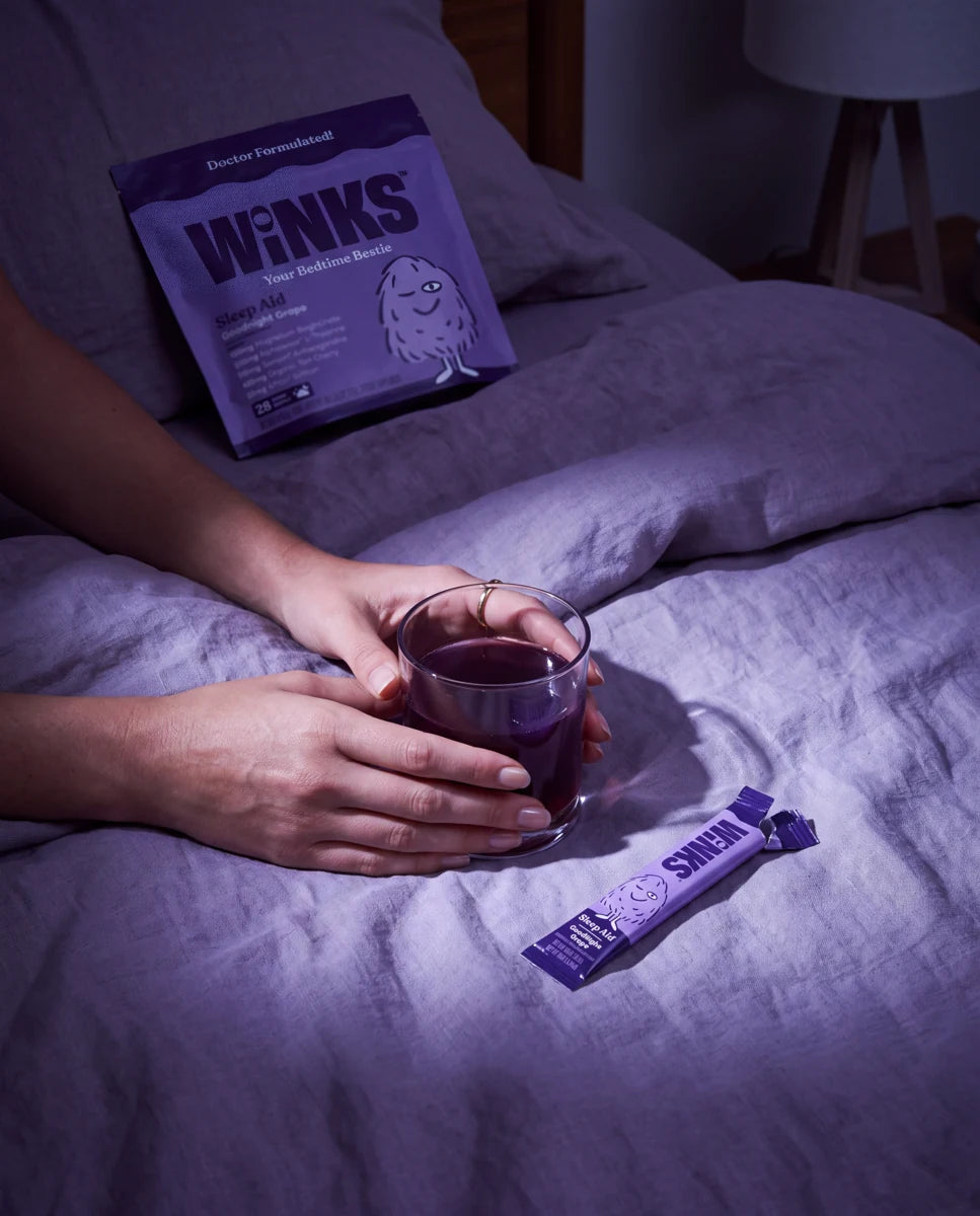 Hands holding a prepared Winks drink on a bed beside a pouch and opened stick.
