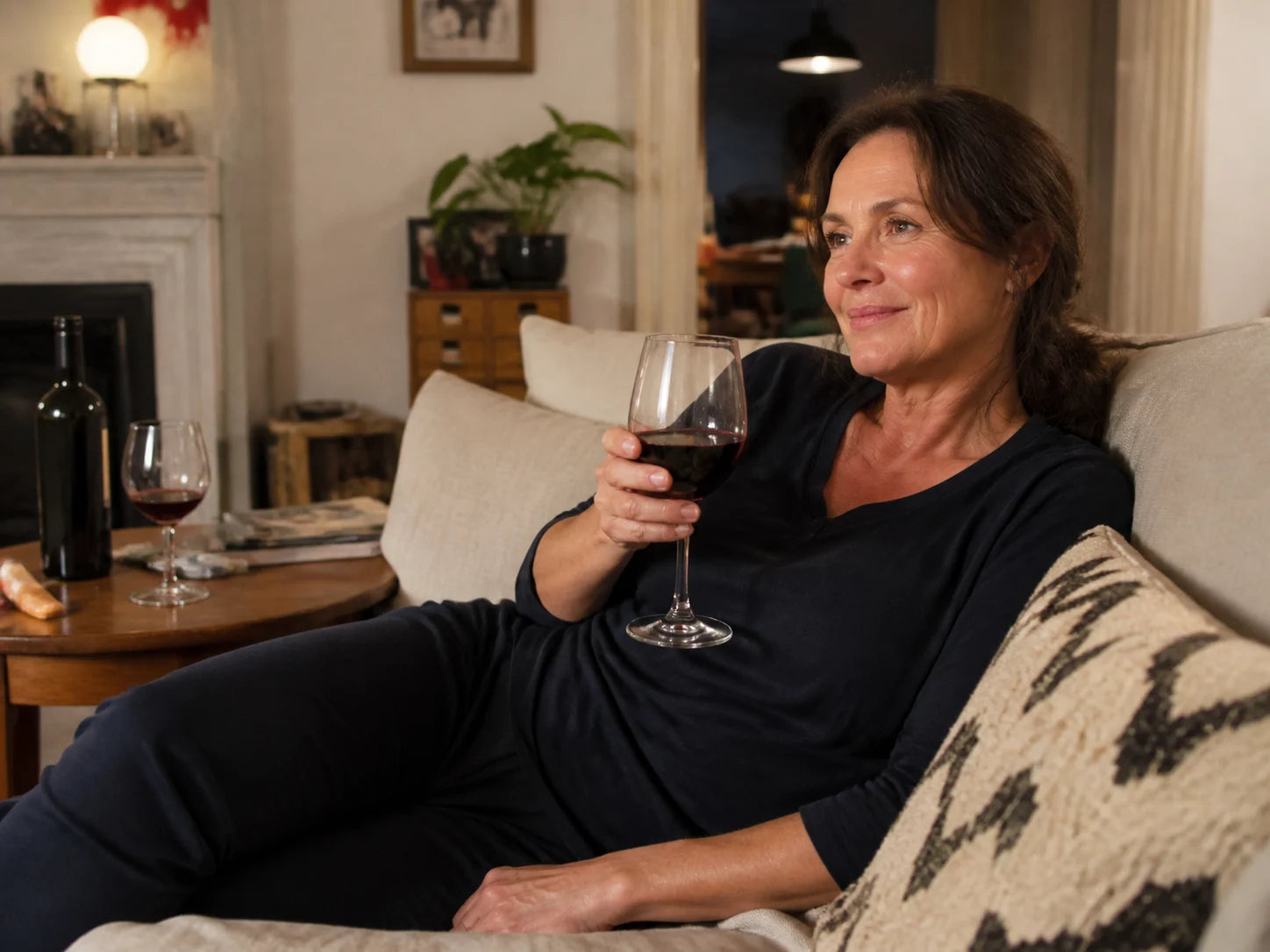 A woman in her late forties holding a glass of red wine on her couch in the evening